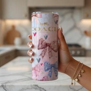May include: A tall, insulated tumbler with a pink and white watercolor design featuring bows, hearts, and the Coach logo. The tumbler is cylindrical and has a gold-colored Coach logo and text. The design includes pink, blue, and green hearts.