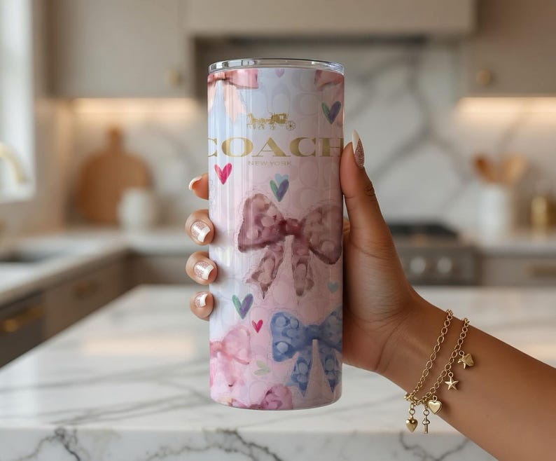 May include: A tall, insulated tumbler with a pink and white watercolor design featuring bows, hearts, and the Coach logo. The tumbler is cylindrical and has a gold-colored Coach logo and text. The design includes pink, blue, and green hearts.