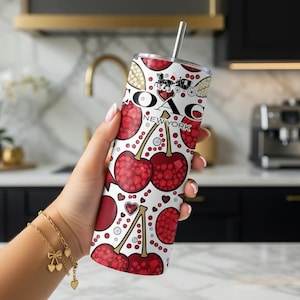 May include: A white tumbler with a cherry and Coach New York pattern. The tumbler has a silver straw and a Coach logo. The design includes red cherries, hearts, and Coach logos. The tumbler is held by a person.