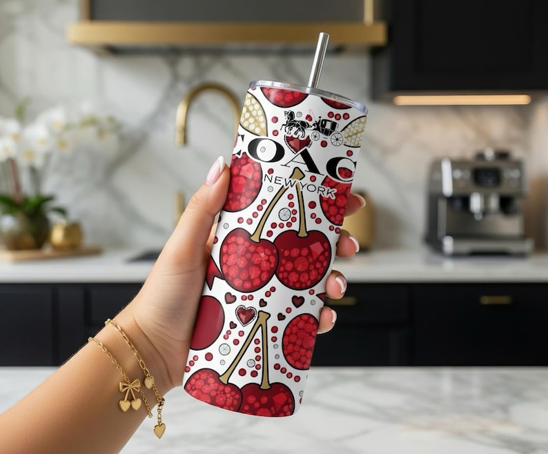 May include: A white tumbler with a cherry and Coach New York pattern. The tumbler has a silver straw and a Coach logo. The design includes red cherries, hearts, and Coach logos. The tumbler is held by a person.