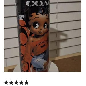 May include: A black tumbler featuring a cartoon image of Betty Boop wearing an orange Coach sweatshirt and ripped blue jeans. The tumbler has the Coach logo and the word "COACH" in white.