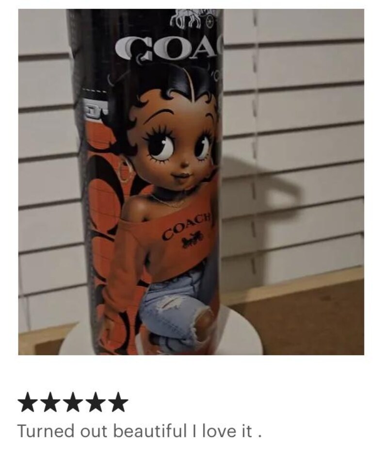 May include: A black tumbler featuring a cartoon image of Betty Boop wearing an orange Coach sweatshirt and ripped blue jeans. The tumbler has the Coach logo and the word "COACH" in white.