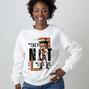 May include: White sweatshirt featuring a cartoon character with a hat and the text "THEY NOT LIKE US". The design includes a red, green, and yellow color scheme, with the word "Juneteenth" on the side.