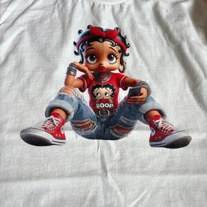 May include: White t-shirt with a cartoon image of Betty Boop wearing a red shirt, blue jeans, and red sneakers. She is holding a cell phone and has a small Betty Boop image on her shirt that says "Boop".