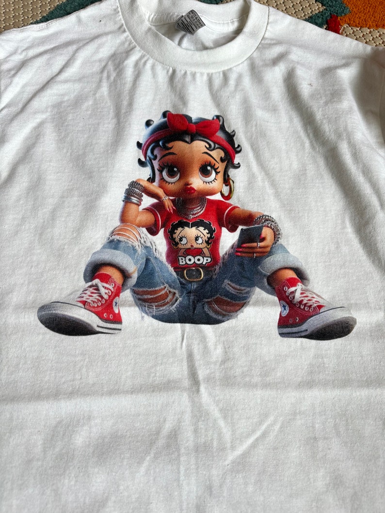 May include: White t-shirt with a cartoon image of Betty Boop wearing a red shirt, blue jeans, and red sneakers. She is holding a cell phone and has a small Betty Boop image on her shirt that says "Boop".