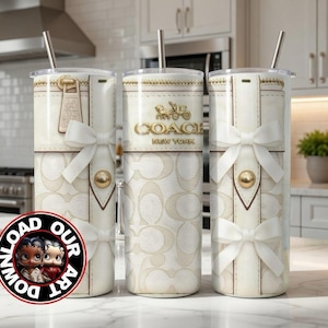 May include: Three Coach New York tumblers with a cream-colored background and a repeating logo pattern. Each tumbler features a white bow, gold accents, and a silver straw. The Coach logo is displayed in gold. The tumblers are on a marble countertop.