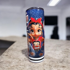 May include: A stainless steel tumbler with a glittery blue background and a cartoon image of Betty Boop. Betty Boop is wearing a red shirt with the word "Boop" and blue jeans. The tumbler has a black and white stripe at the bottom.