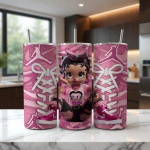 May include: Pink tumbler with a Betty Boop doll and a pink and white sneaker design. The tumbler features a silver straw and a pink and white shoelace design. The Betty Boop doll is wearing a pink shirt and black pants.