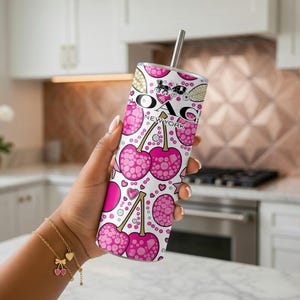 May include: A white tumbler with a pink cherry pattern and the text "OAG NEW YORK". The tumbler has a silver straw and is held in a hand. A gold bracelet with cherry charms is also visible.