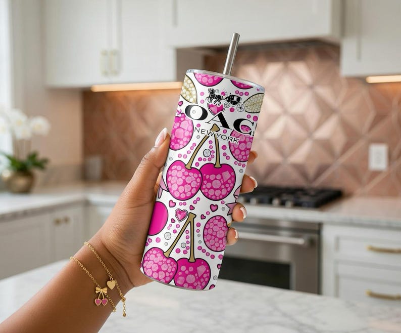 May include: A white tumbler with a pink cherry pattern and the text "OAG NEW YORK". The tumbler has a silver straw and is held in a hand. A gold bracelet with cherry charms is also visible.