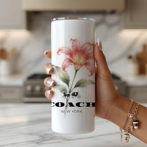 May include: White tumbler featuring a watercolor-style pink lily and green leaves. The tumbler has the Coach New York logo and a horse-drawn carriage graphic. The tumbler is held by a person.