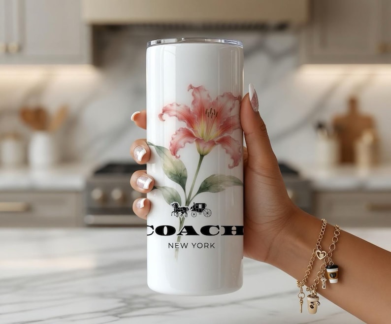 May include: White tumbler featuring a watercolor-style pink lily and green leaves. The tumbler has the Coach New York logo and a horse-drawn carriage graphic. The tumbler is held by a person.