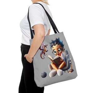 May include: A gray tote bag with a cartoon image of a girl reading a book with a dragon beside her. The girl is wearing glasses and a white shirt. The bag has black handles.