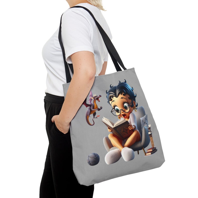 May include: A gray tote bag with a cartoon image of a girl reading a book with a dragon beside her. The girl is wearing glasses and a white shirt. The bag has black handles.