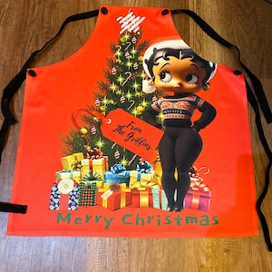 Może przedstawiać: Czerwony świąteczny fartuch z postacią z kresk&oacute;wki Betty Boop w czapce i swetrze Świętego Mikołaja, obok udekorowanej choinki i zapakowanych prezent&oacute;w. Fartuch ma czarne wiązania i napis "Merry Christmas" na dole.
