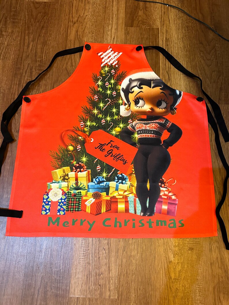 Może przedstawiać: Czerwony świąteczny fartuch z postacią z kresk&oacute;wki Betty Boop w czapce i swetrze Świętego Mikołaja, obok udekorowanej choinki i zapakowanych prezent&oacute;w. Fartuch ma czarne wiązania i napis "Merry Christmas" na dole.
