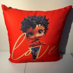 May include: Red pillow with a cartoon image of a woman with black hair and big eyes. The woman is wearing a red shirt, blue jeans, and is making a heart shape with her hands. The text "Love Yourself" is written in gold lettering below the image.