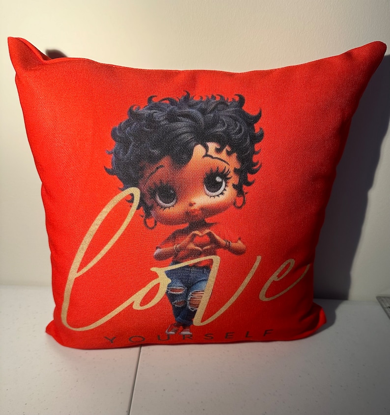 May include: Red pillow with a cartoon image of a woman with black hair and big eyes. The woman is wearing a red shirt, blue jeans, and is making a heart shape with her hands. The text "Love Yourself" is written in gold lettering below the image.