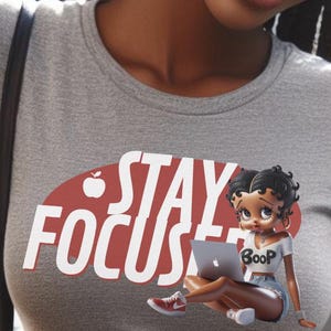 May include: Gray t-shirt with the words "STAY FOCUS" in white letters over a red oval. A cartoon figure of a woman wearing a white crop top with "Boop" on it, denim shorts, and red sneakers is holding a laptop.