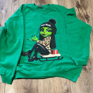 May include: Green sweatshirt with a graphic of a green-skinned woman wearing a leopard print outfit and a hat that says "Hum Bug". She is sitting on a red sled.
