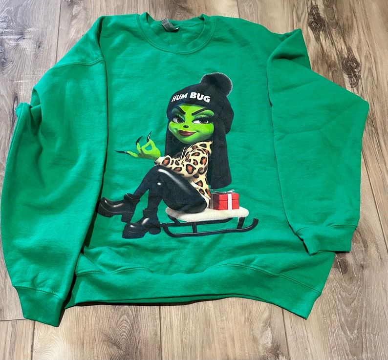 May include: Green sweatshirt with a graphic of a green-skinned woman wearing a leopard print outfit and a hat that says "Hum Bug". She is sitting on a red sled.