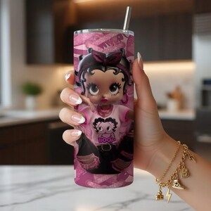 May include: A pink tumbler with a cartoon image of Betty Boop. The tumbler has a stainless steel straw and a repeating pattern of pink bows and the word "Boop". The cartoon character is wearing a pink shirt with the word "Boop".