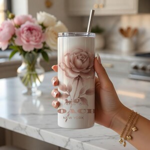 May include: A white tumbler with a rose design and a silver straw. The tumbler features the Coach New York logo and a floral illustration in shades of pink and brown. A gold bracelet with charms is visible.