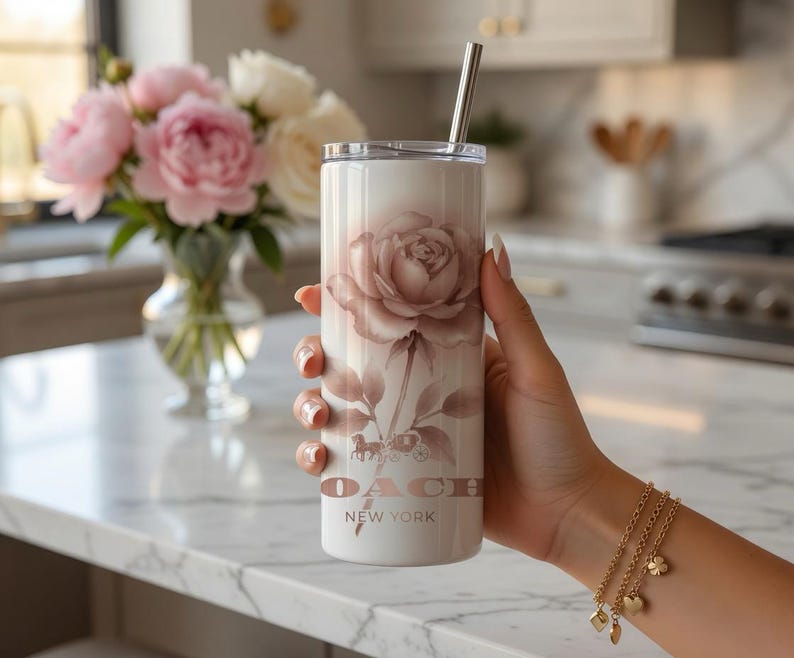 May include: A white tumbler with a rose design and a silver straw. The tumbler features the Coach New York logo and a floral illustration in shades of pink and brown. A gold bracelet with charms is visible.