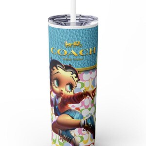 May include: A tall, white tumbler with a lid and straw. The tumbler features a cartoon image of Betty Boop in a red jacket and blue shorts. The top section has a blue leather-like texture with the gold "COACH NEW YORK" logo. The background has a repeating pattern.
