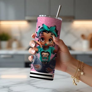 May include: A pink glitter tumbler with a cartoon character wearing a teal bow and a shirt that says "BOOP". The tumbler has a silver straw and a black and white striped base. A gold charm bracelet is visible.
