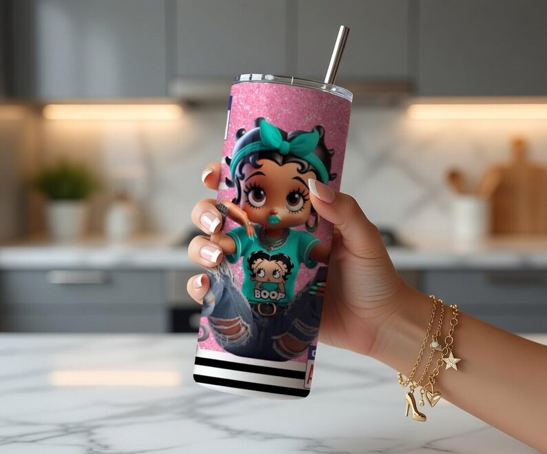 May include: A pink glitter tumbler with a cartoon character wearing a teal bow and a shirt that says "BOOP". The tumbler has a silver straw and a black and white striped base. A gold charm bracelet is visible.