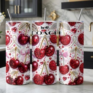 May include: Three stainless steel tumblers with a white background, adorned with a pattern of red cherries, hearts, and diamond accents. The Coach New York logo is visible on one tumbler. Each tumbler has a silver straw.