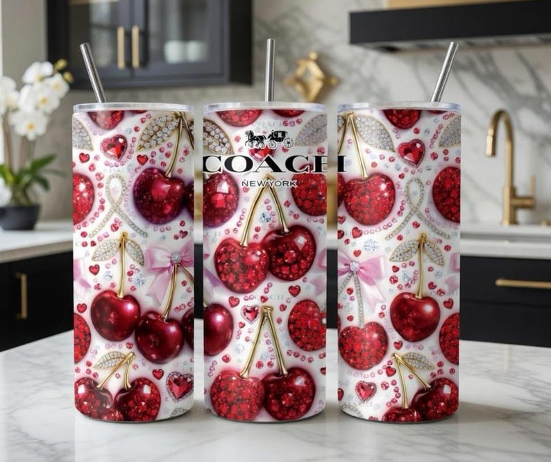 May include: Three stainless steel tumblers with a white background, adorned with a pattern of red cherries, hearts, and diamond accents. The Coach New York logo is visible on one tumbler. Each tumbler has a silver straw.