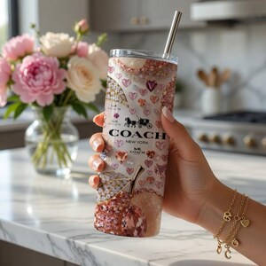 May include: A Coach New York tumbler with a silver straw, featuring a design of hearts, gems, and a cherry. The tumbler has the Coach logo and a horse-drawn carriage emblem. The background includes a bouquet of pink and white flowers.