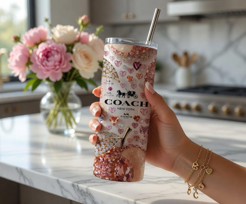 May include: A Coach New York tumbler with a silver straw, featuring a design of hearts, gems, and a cherry. The tumbler has the Coach logo and a horse-drawn carriage emblem. The background includes a bouquet of pink and white flowers.