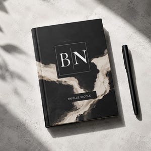 以下が含まれることがあります： 黒とベージュのマーブル模様の表紙を持つハードカバーのジャーナル。表紙には、文字「BN」が入った白い正方形のフレームと、「BRIELLE NICOLE」という白い文字が書かれた黒い長方形のラベルがあります。横には黒いペンが置かれています。