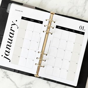 May include: Open planner with a January calendar page. The calendar is white with black text and a gold-colored binder. The word "january" is written vertically on the left side of the page.