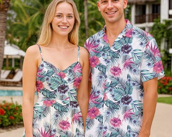 May include: A matching set of tropical-themed clothing. A woman wears a white sundress with a floral pattern of pink, purple, and teal. A man wears a short-sleeved button-down shirt with the same floral design, paired with white shorts. Palm trees and a resort are in the background.