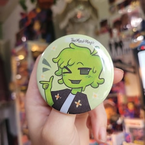 May include: A round, light green button with a cartoon character design. The character is green with a black shirt and white undershirt. The text "The Most Mayo" is at the top, with small white flowers around the edge.
