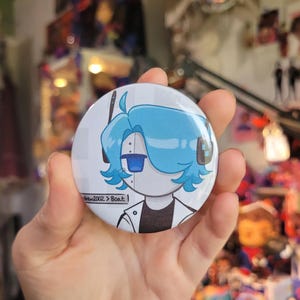 May include: A round, white button featuring a cartoon robot character with blue hair and a blue eye. The robot is wearing a white jacket with a black shirt underneath. The button has the text "andrew2002 > Boat:" printed on it.