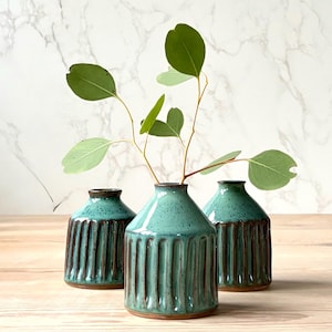 May include: Three teal ceramic vases with vertical grooves and dark brown rims. Each vase holds a sprig of green eucalyptus leaves. The vases are arranged on a light wooden surface against a marble background.