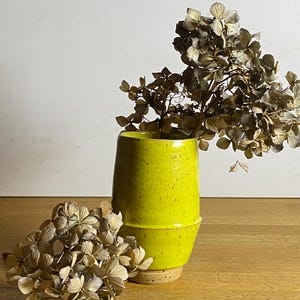 May include: A yellow ceramic vase with a speckled glaze, filled with dried white hydrangeas. The vase is sitting on a wooden surface.