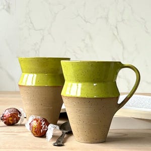 May include: Two handmade ceramic mugs with a light green glazed top and a speckled tan base. The mugs have a curved handle. Two wrapped chocolate candies and a spoon are on the wooden surface. The mugs are approximately 10 cm tall.