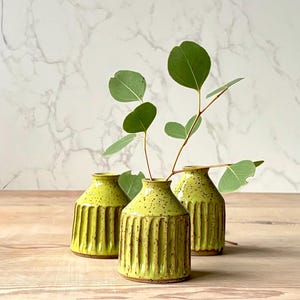 May include: Three small, lime green ceramic vases with vertical ribbed detailing. Each vase holds a sprig of green eucalyptus leaves. The vases are arranged on a light wood surface against a white marble-patterned background.