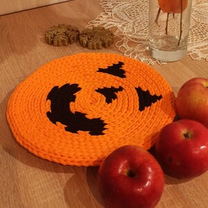Puede incluir: Posavasos de punto de ganchillo naranja en forma de calabaza con un diseño de cara de calabaza negra. Tres manzanas rojas están sobre una superficie de madera junto al posavasos.