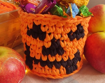 Può includere: Un cestino all'uncinetto arancione e nero a forma di zucca, pieno di caramelle incartate. Il cestino è circondato da mele rosse e lucine, creando un'atmosfera festosa di Halloween.