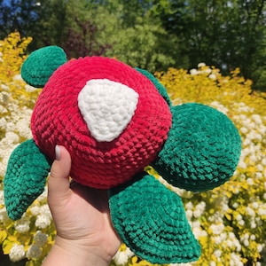 Puede incluir: Un peluche de tortuga hecho a mano con ganchillo. La tortuga tiene un cuerpo rojo con un parche en forma de corazón blanco y aletas y cabeza verdes. El peluche está hecho de hilo suave y texturizado.