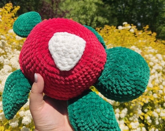 May include: A handmade crochet turtle plushie. The turtle features a red body with a white heart-shaped patch and green flippers and head. The plushie is made of soft, textured yarn.