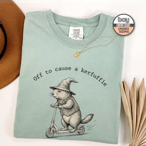 May include: A light green unisex t-shirt with the phrase "Off to cause a kerfuffle" and a graphic of a groundhog riding a scooter and wearing a wizard hat. A gold necklace is draped across the shirt. The shirt has a Comfort Colors tag.