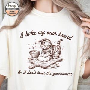 May include: A white t-shirt with a brown graphic of a cat kneading dough on a cutting board. The text "I bake my own bread & I don't trust the government" is printed on the shirt.